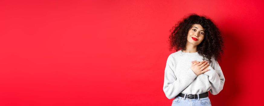 Tender young woman with curly hairstyle, holding hands on heart and smiling with grateful expression, say thank you, standing on red background.