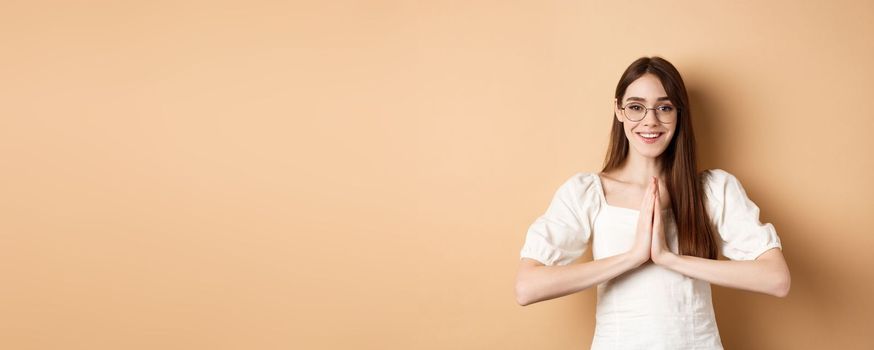 Smiling girl in glasses say thank you, show namaste gesture, express gratitude, standing pleased on beige background.