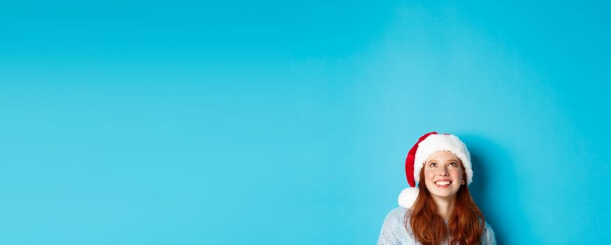 Winter holidays and Christmas eve concept. Head of cute redhead girl in santa hat, appear from bottom and looking up at copy space, staring logo, standing over blue background.