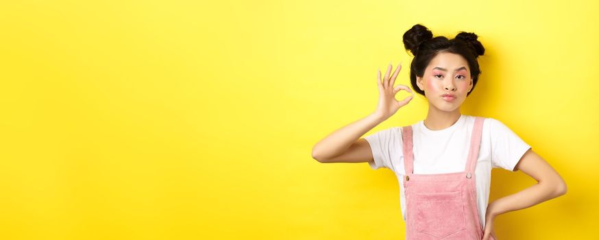 Sassy teenage asian girl with bright pink makeup, showing okay sign and nod in approval, approve product with serious face, not bad gesture, standing against yellow background.