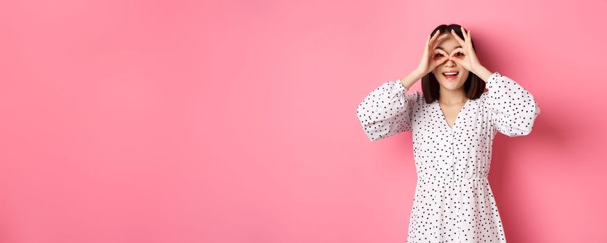Carefree young asian woman looking through hand binoculars at camera, staring at discounts, standing over pink background.
