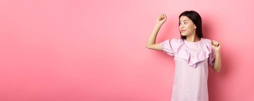 Carefree asian girl dancing, raising hand up and looking left at logo, standing in romantic dress against pink background.