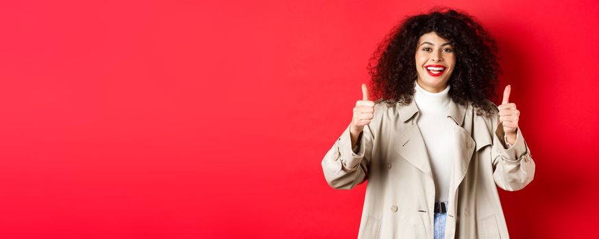 Portrait of young woman looking happy, wearing trench coat and showing thumbs-up, say yes, approve and praise something good, red background.