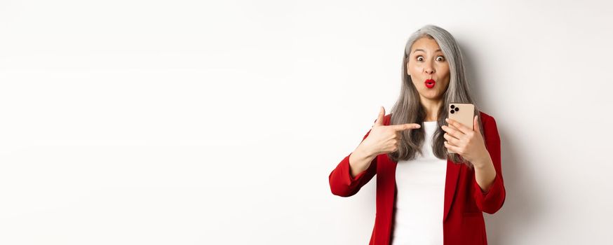 Amazed korean lady in red blazer pointing finger at mobile phone, looking curious at camera, white background.