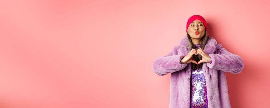 Romance and valentines day. Happy asian senior woman showing heart sign, I love you gesture, pucker lips for kiss, standing over pink background.