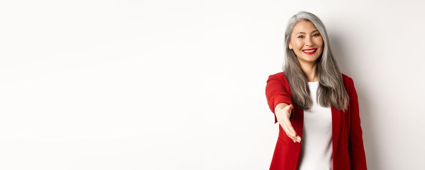 Professional asian businesswoman with grey hair, saying hello, stretch out hand for handshake and smiling, standing over white background.