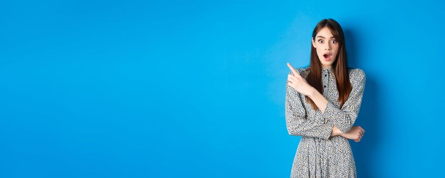 Excited pretty woman in dress pointing at upper left corner, look with disbelief and amazement, checking out cool promo, blue background.