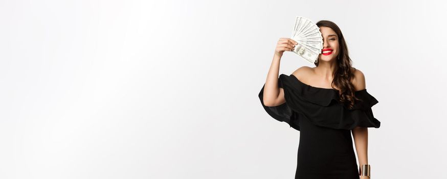 Fashion and shopping concept. Happy young woman in black dress, with red lips, holding money and smiling satisfied, standing over white background.