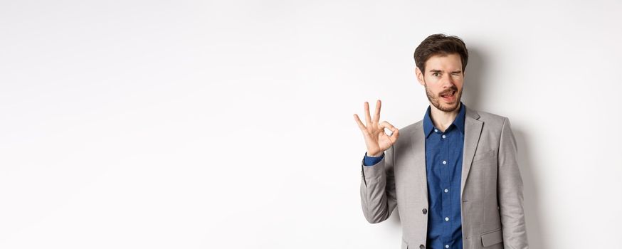 All good. Confident male ceo manager showing okay sign and winking, everything under control, praising nice work, standing on white background.