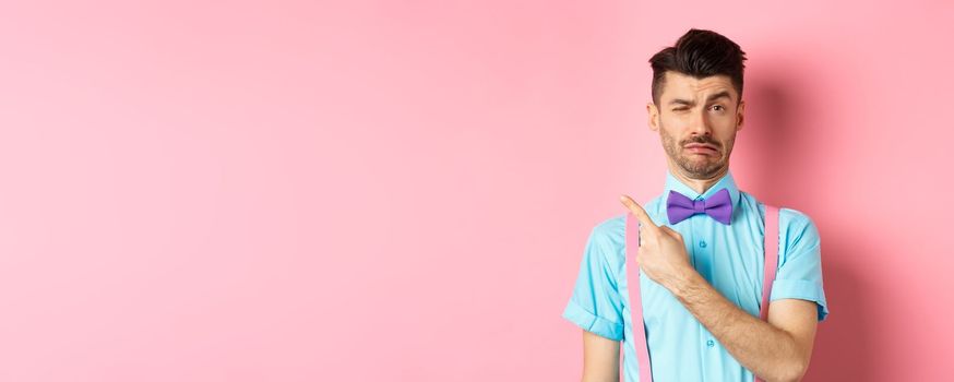 Childish young man acting sad and fake crying while pointing finger upper left corner, complaining on something, standing over pink background.