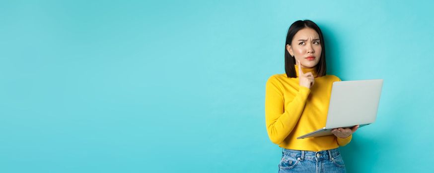 Pensive asian woman working on laptop, thinking and looking away, making decision, standing against blue background.