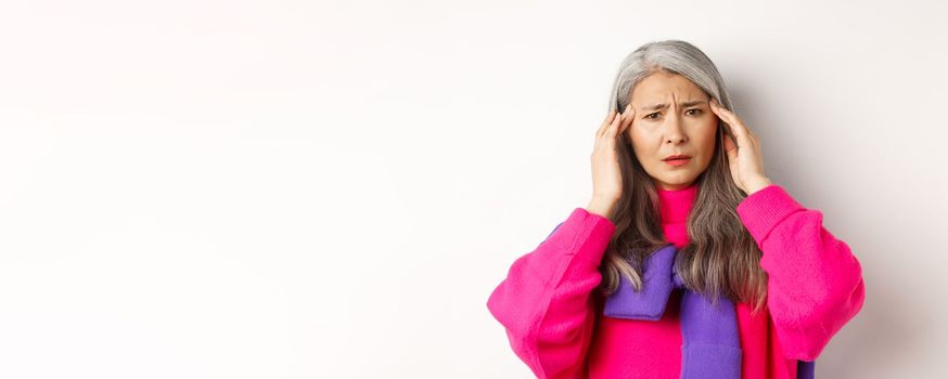 Close-up of fashionable asian senior woman feeling unwell, touching head and grimacing from headache, complain on migraine, white background.