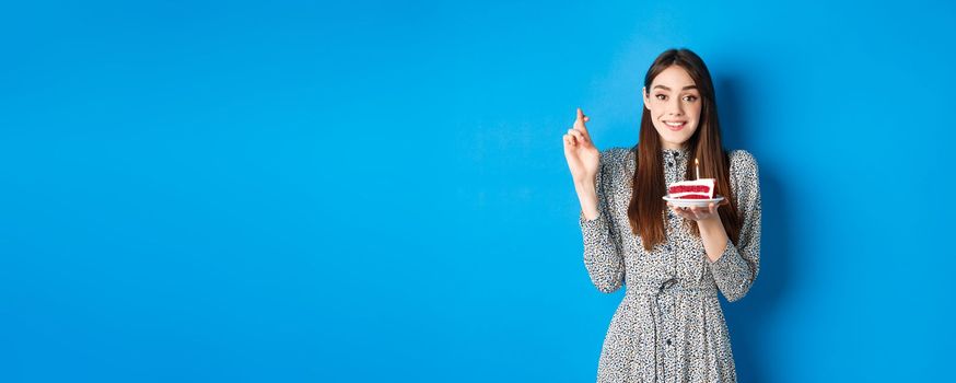 Hopeful birthday girl making wish on cake, cross fingers for good luck, standing in stylish dress against blue background.