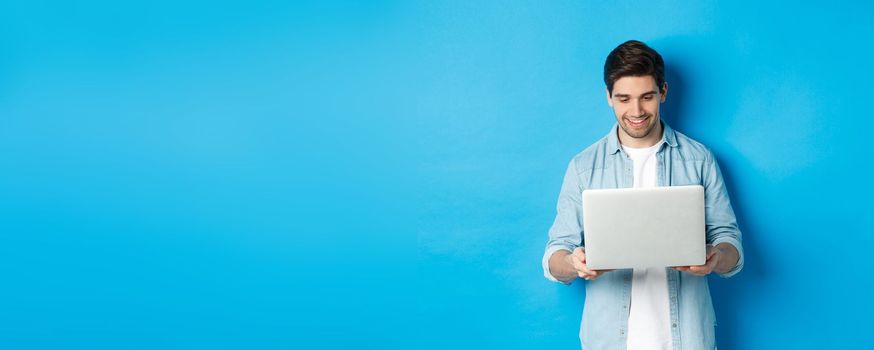 Handsome man working on laptop, smiling and looking at screen satisfied, standing against blue background.