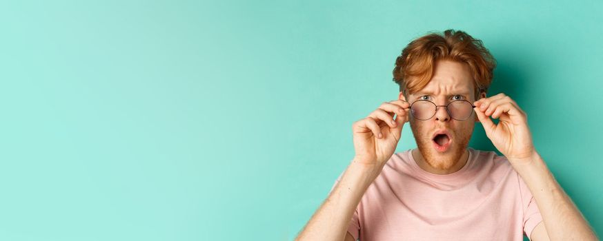 Close up portrait of redhead guy take-off glasses and gasping startled, looking shocked at camera, standing over turquoise background.