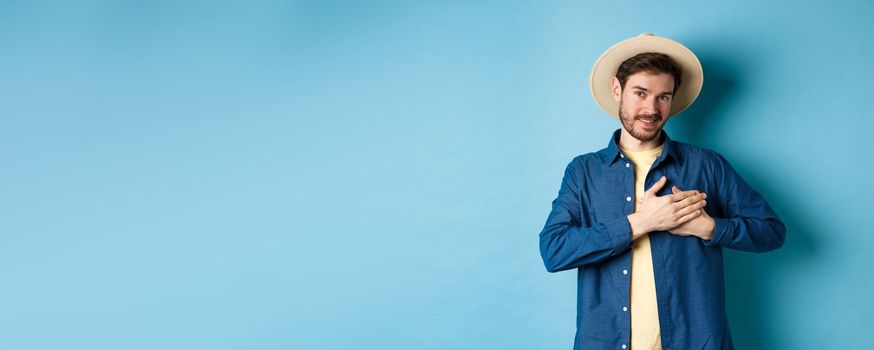 Handsome traveler in straw hat, holding hands on heart and smiling, showing thank you gesture, keep memories about vacation in soul, standing on blue background.