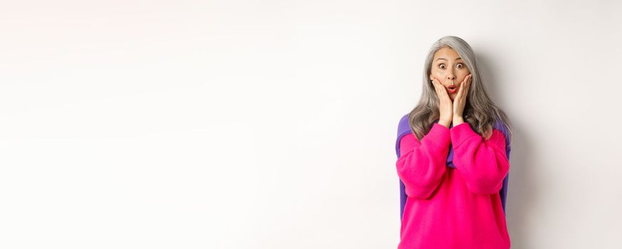 Surprised asian senior woman gasping amazed, holding hands on face and staring at camera impressed, standing over white background.