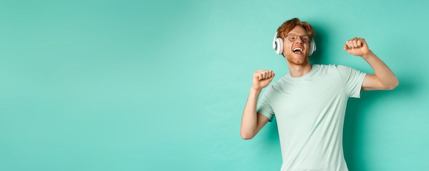 Lifestyle concept. Carefree guy with red hair, dancing and having fun, listening music in headphones, standing over turquoise background.