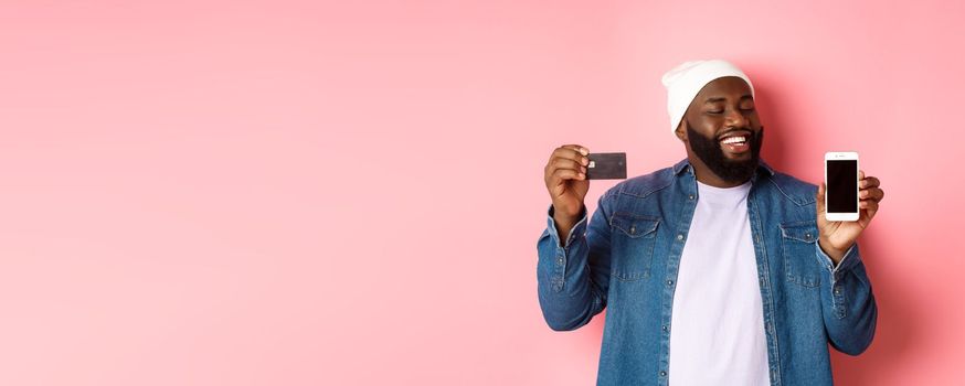Online shopping. Satisfied Black man nod in approval, smiling and looking at phone, showing credit card and smartphone screen, standing over pink background.