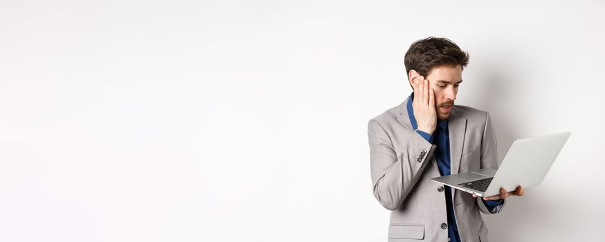 Shocked and worried businessman making mistake on laptop, looking at computer upset, standing on white background.