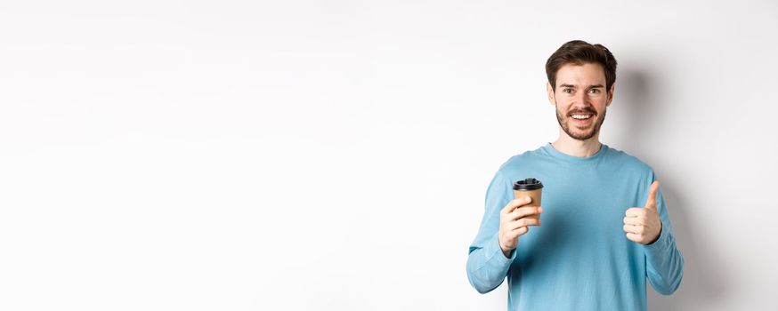 Cheerful young man drinking good coffee, holding paper cup and showing thumb up, recommending cafe shop, standing over white background.