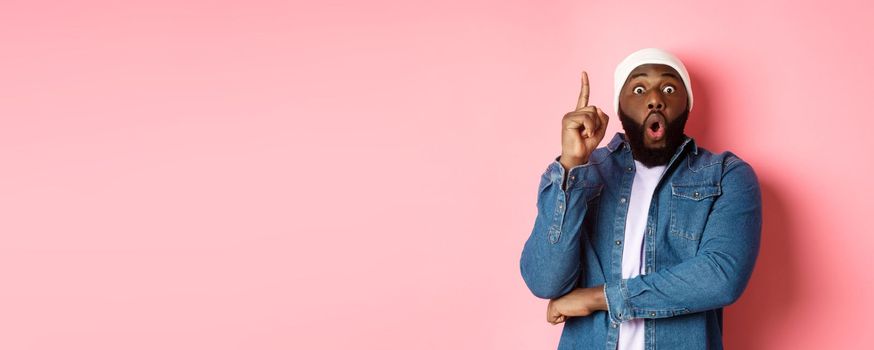 Handsome hipster Black guy having an idea, raising finger up and saying suggestion, got solution, standing over pink background.