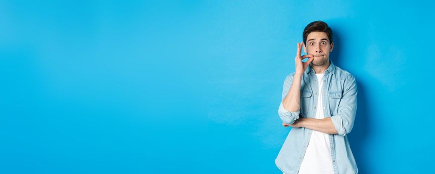 Adult man zipping mouth, promise keep secret, making a seal on lips and standing over blue background.