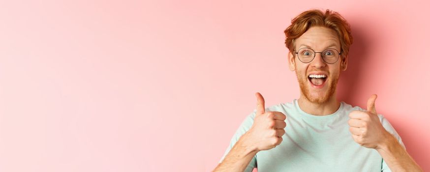 Face of happy redhead man in glasses and t-shirt, showing thumbs-up and looking excited, approve and praise cool promotion, standing over pink background.