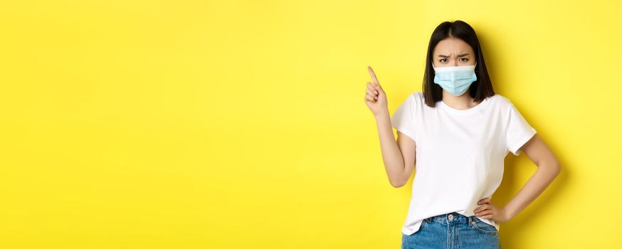 Covid, health care and pandemic concept. Asian female model in medical mask and white t-shirt pointing finger at upper left corner logo, showing promotion, yellow background.