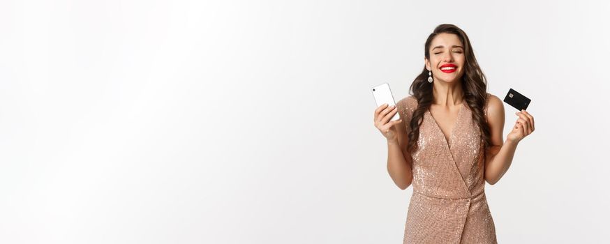 Online shopping and holidays concept. Satisfied and happy woman in elegant dress smiling, using credit card and mobile phone, standing over white background.