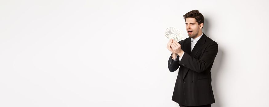 Image of confused businessman looking at fake money, standing over white background in black suit.