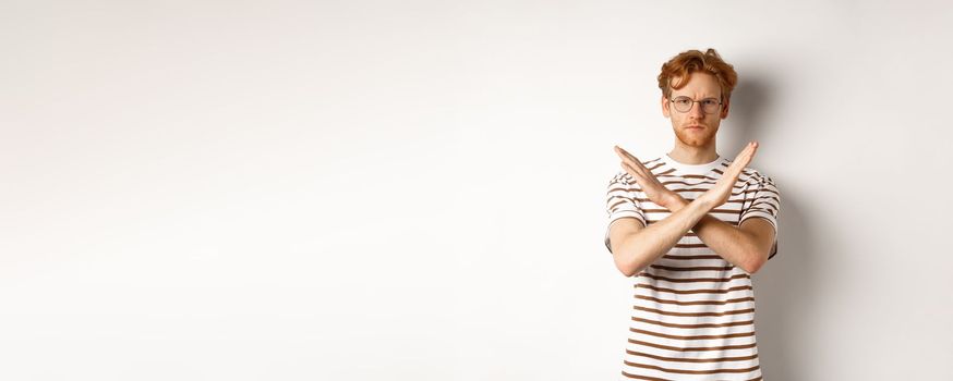 Serious young man with ginger hair frowning, showing stop gesture, making cross to prohibit something bad, disagree with you, standing over white background.