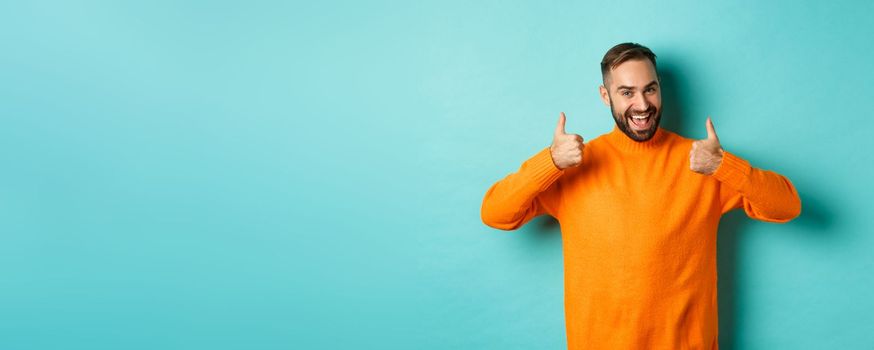 Handsome man expressing support, showing thumb up, encourage you, praise excellent work, approve and agree, standing over light blue background.