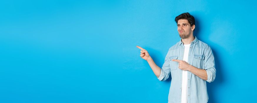 Doubtful adult man pointing fingers left at promotion and looking unsure, grimacing disappointed, standing against blue background.