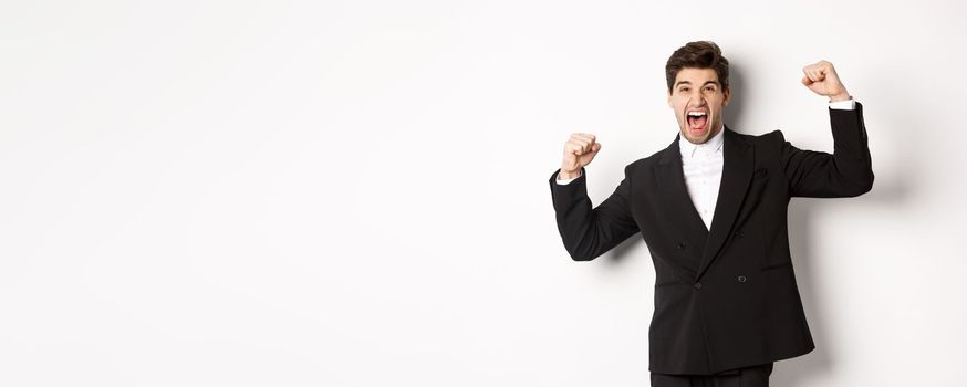 Portrait of successful businessman in black suit, become champion, raising hands up and shouting yes, triumphing and celebrating victory, standing against white background.