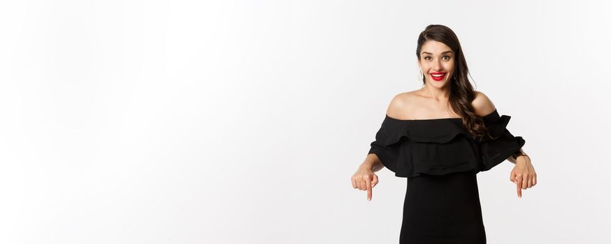 Fashion and beauty. Elegant woman in black dress pointing fingers down, showing promo and smiling, standing over white background.