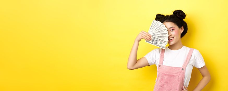 Shopping. Beautiful asian female model with glamour makeup, cover half of face with money dollar bills, smiling sensual at camera, yellow background.