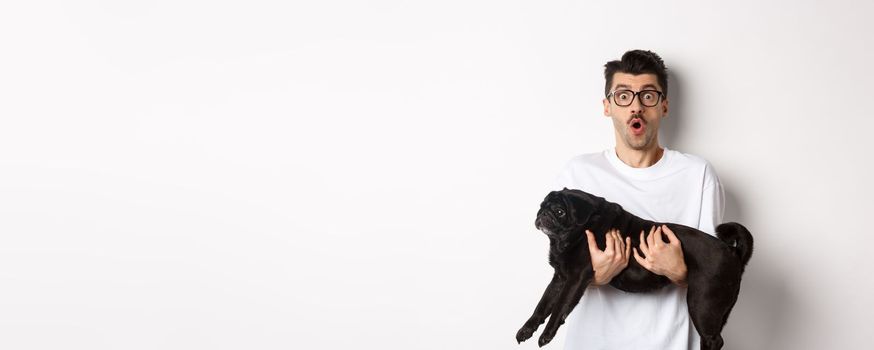 Surprised young man in glasses holding cute black pug, dog owner staring at camera with impressed face, saying wow, standing over white background.