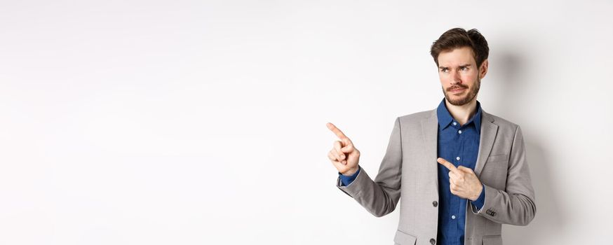 Displeased businessman in suit pointing and looking left with skeptical smirk, dislike banner, disapprove promo offer, standing on white background.