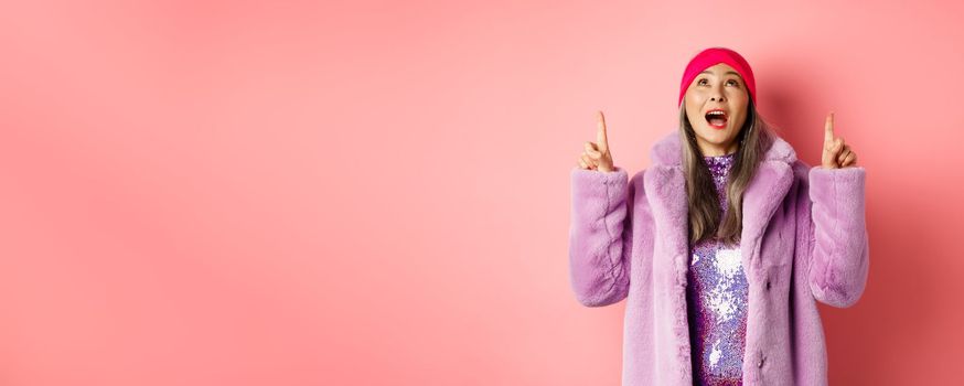 Mature asian hipster woman pointing fingers up, looking at top promo with amazement, checking out special deal, standing over pink background.