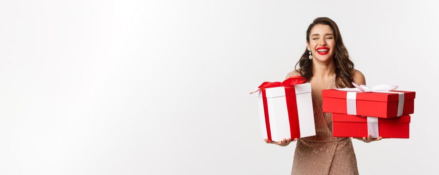 Holidays, celebration concept. Beautiful caucasian woman in elegant dress holding Christmas presents and smiling happy, standing over white background.