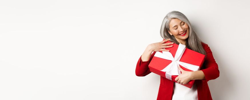 Valentine day concept. Beautiful asian senior woman hugging box with gift, smiling and looking happy, receiving present on holiday, white background.