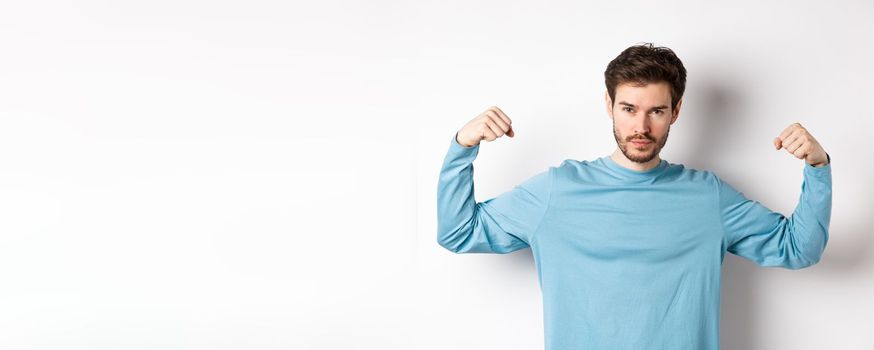 Confident and strong macho man flexing biceps, showing strength in muscles after gym workout, standing over white background.