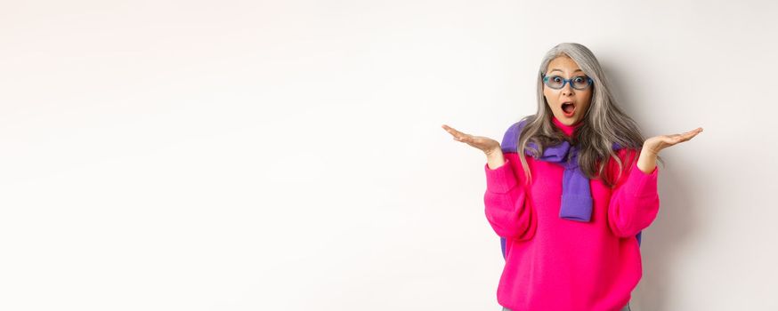 Fashionable asian senior woman in sunglasses gasping amazed, spread hands sideways and staring surprised at camera, white background.