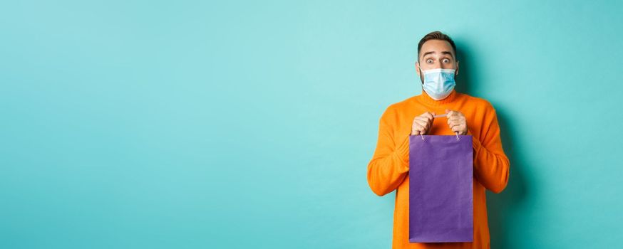 Covid-19, pandemic and lifestyle concept. Surprised man open shopping bag and looking amazed, receiving gift on holiday, standing over turquoise background.