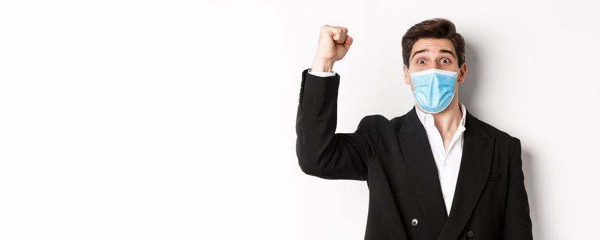 Concept of covid-19, business and social distancing. Close-up of excited businessman in medical mask and suit, rejoicing, achieving goal and triumphing, white background.