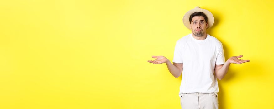 Concept of tourism and summer. Confused male tourist shrugging, looking indecisive, standing against yellow background.