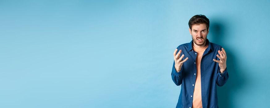 Annoyed and angry man raising hands and clench teeth outraged, stare pissed-off at camera, going to kill someone, standing on blue background.