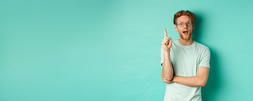 Excited young man with ginger hair in glasses, raising index finger, pitching an idea, standing over mint background. Copy space