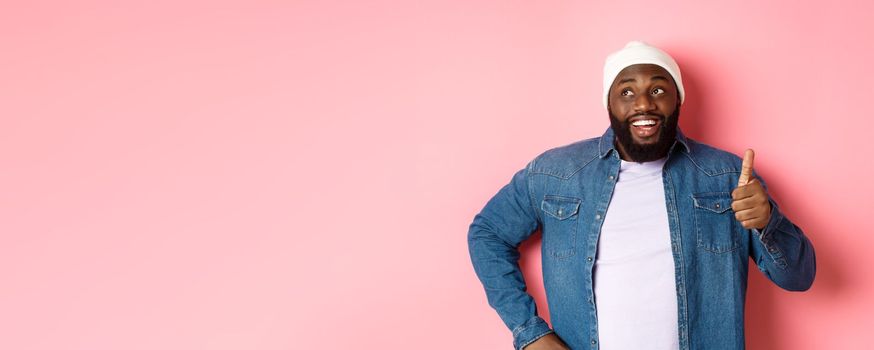 Happy Black man showing thumb-up, looking at upper left corner thoughtful, standing over pink background.